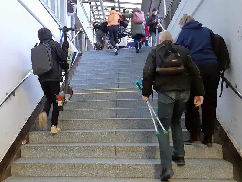 Foto: Lüneburg Barrierefrei. Kein Fahrstuhl: Der Schnappschuss zeigt, wie viele betroffen sind. Rechts ein Herr, der mit Krücken unterwegs ist, links trägt jemand seinen E-Roller. Der Kinderwagen, den drei Personen hochtragen, ist schon fast oben angekommen.