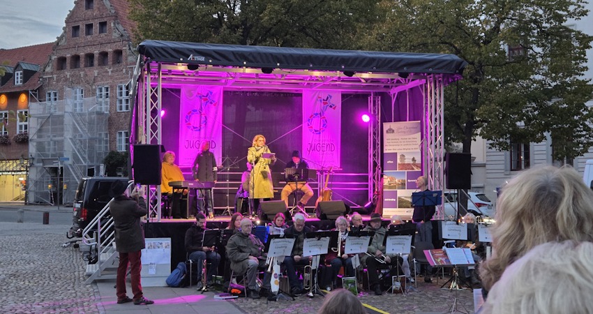 Foto: Lüne-Blog. "Deutschland singt" - 3. Oktober 2025. OB Claudia Kalisch begrüßt die Menschen auf dem Marktplatz. Im Vordergrund: Posaunenchor des Kirchenkreises Lüneburg. Rechts auf der Bühne Klaus Stehr von Noordlücht.