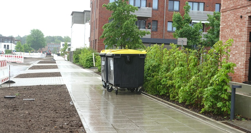 Foto: Uwe Wenk, 11.05.2026. Gehweg an der Lübecker Straße. 
Positiv: Eine angenehm begeh- und befahrbare Oberfläche für Gehhilfen.
Aber: Die Standard-Gehwegbreite beträgt 2,50 Meter. Dann können zwei Menschen bequem nebeneinander und aneinander vorbei gehen. Obwohl hier genügend Platz nach links wäre, ist der Gehweg hier aber nur 2 Meter breit. Zudem ist er zugestellt mit Müllcontainern. Für Fußgänger bleiben nur noch etwa 1,20 Meter - für den Rollstuhl zu knapp.