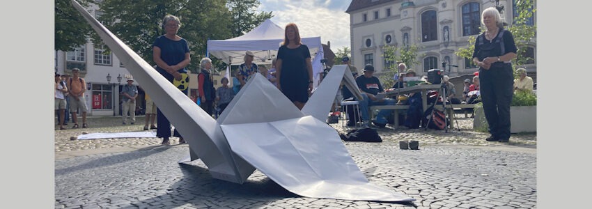 Foto: Dirk Werner. Anlässlich des Gedenktags zu den Atombombenabwürfen über Hiroshima und Nagasaki am 09.08.2025 halten Mitglieder des Friedensbündnisse eine Mahnwache auf dem Marktplatz Lüneburg. Vor ihnen liegt ein gefalteter Kranich aus Papier als Friedenssymbol.